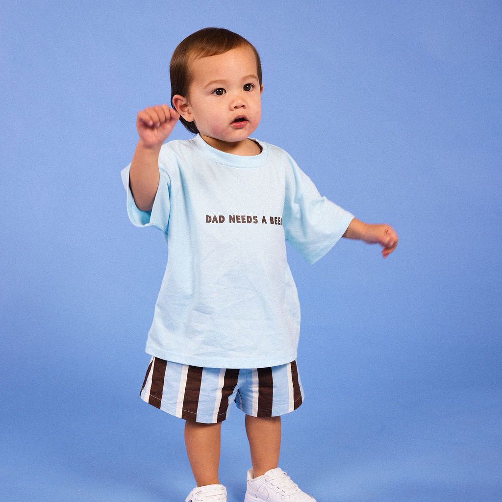 A toddler standing against a solid blue studio background, wearing a light blue graphic T-shirt with the text 'DAD NEEDS A BEER', brown and white striped shorts, and white sneakers.