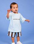A toddler standing against a solid blue studio background, wearing a light blue graphic T-shirt with the text 'DAD NEEDS A BEER', brown and white striped shorts, and white sneakers.