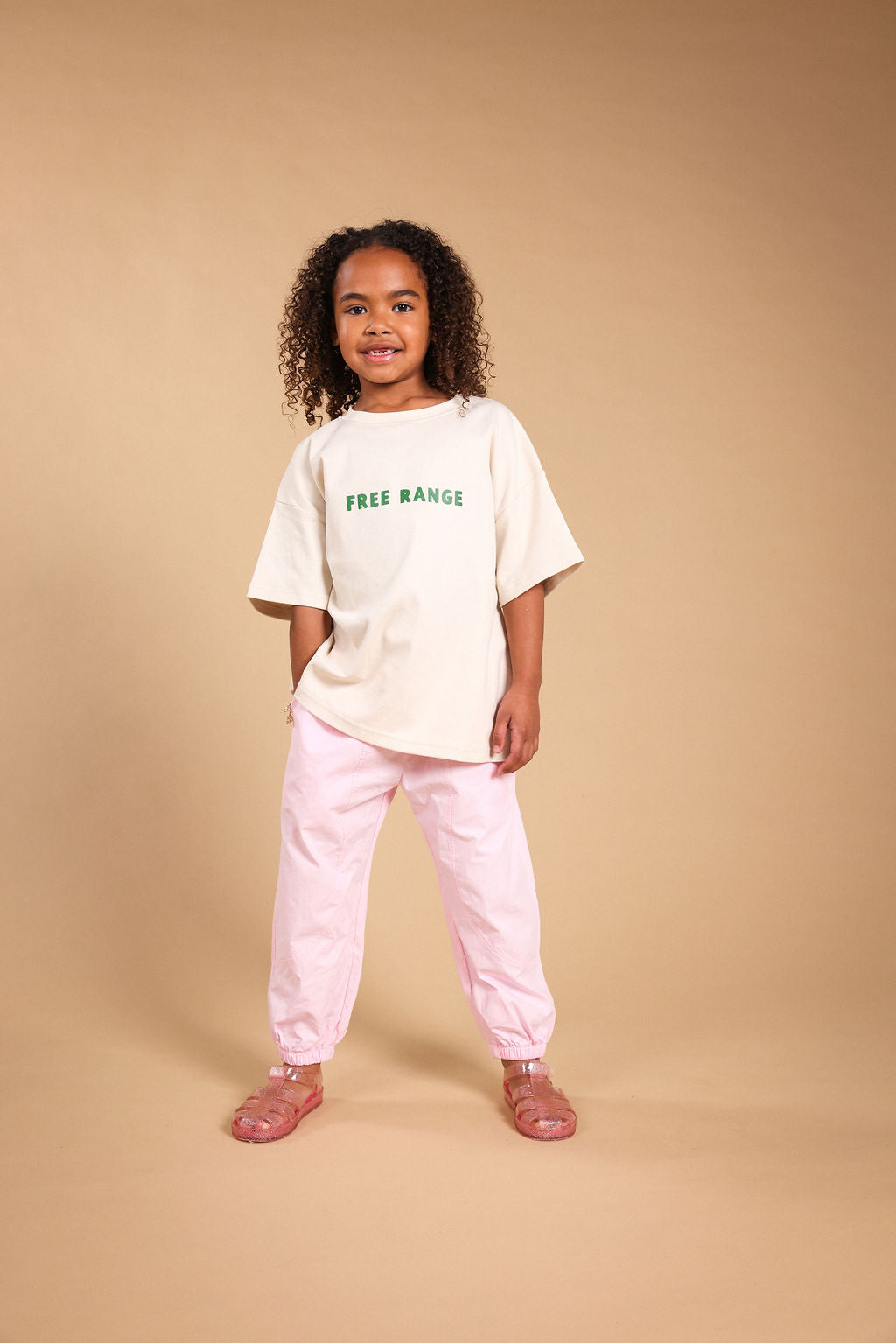 A young girl with curly dark hair stands against a solid beige studio background, wearing an oversized off-white t-shirt with green 'FREE RANGE' text, light pink pants, and pink sandals.