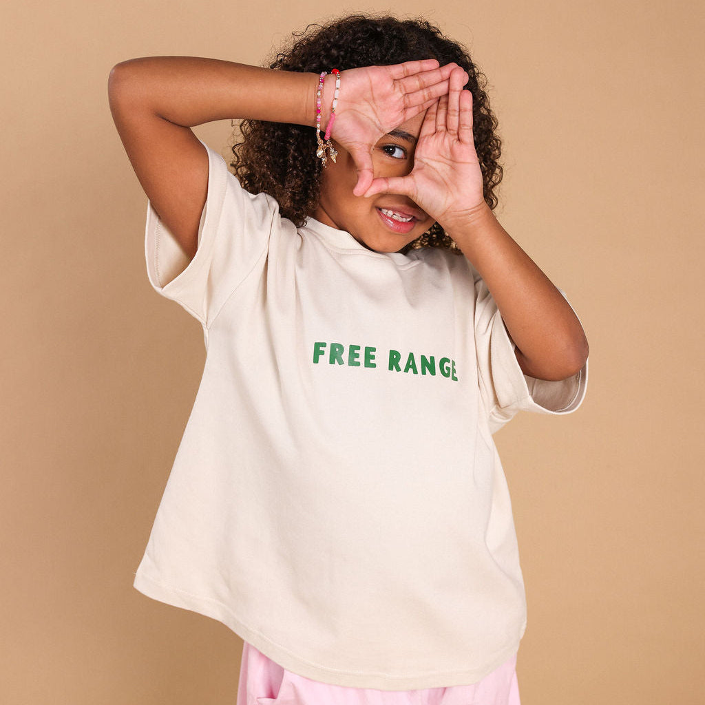 A child wearing a cream-colored oversized t-shirt with green 'FREE RANGE.' text on the chest, paired with light pink pants, posing against a solid beige background.