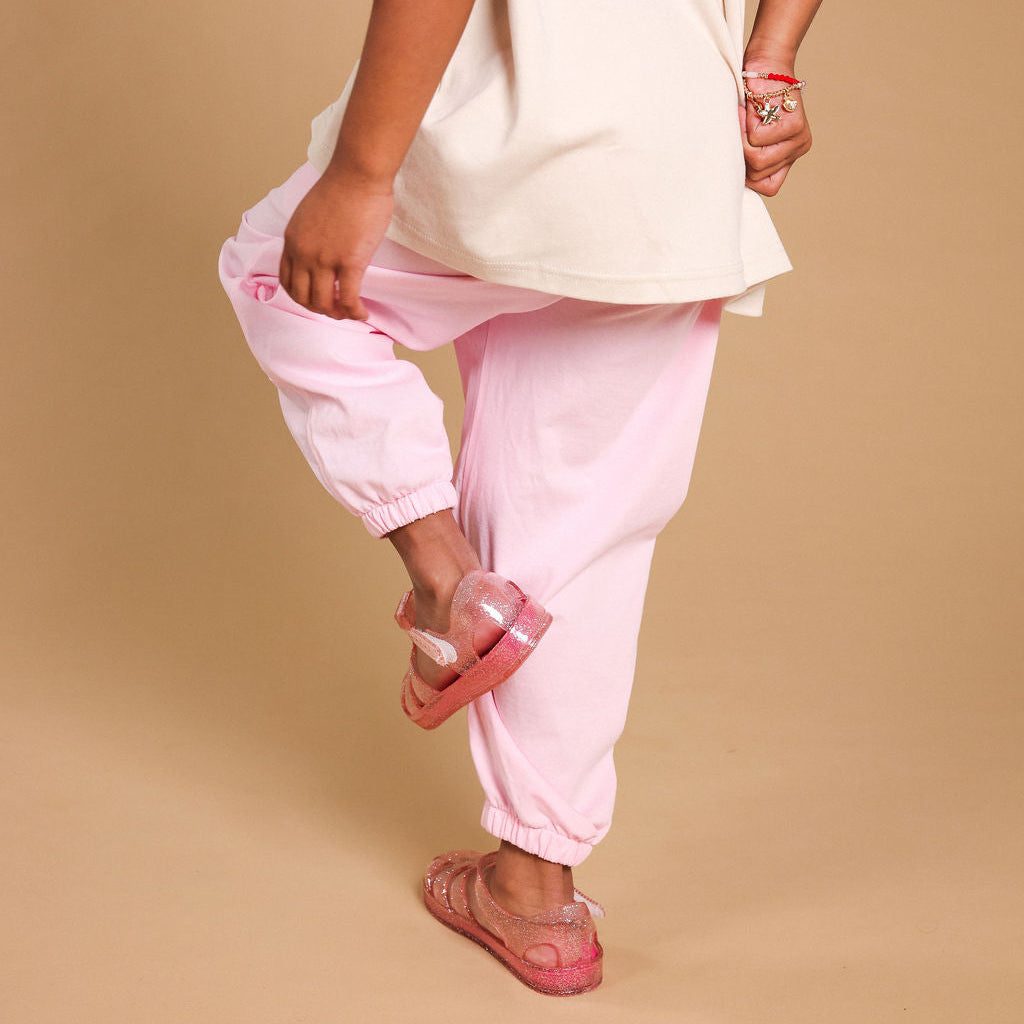 A child wearing an oversized cream t-shirt and light pink jogger pants, standing against a solid beige background.
