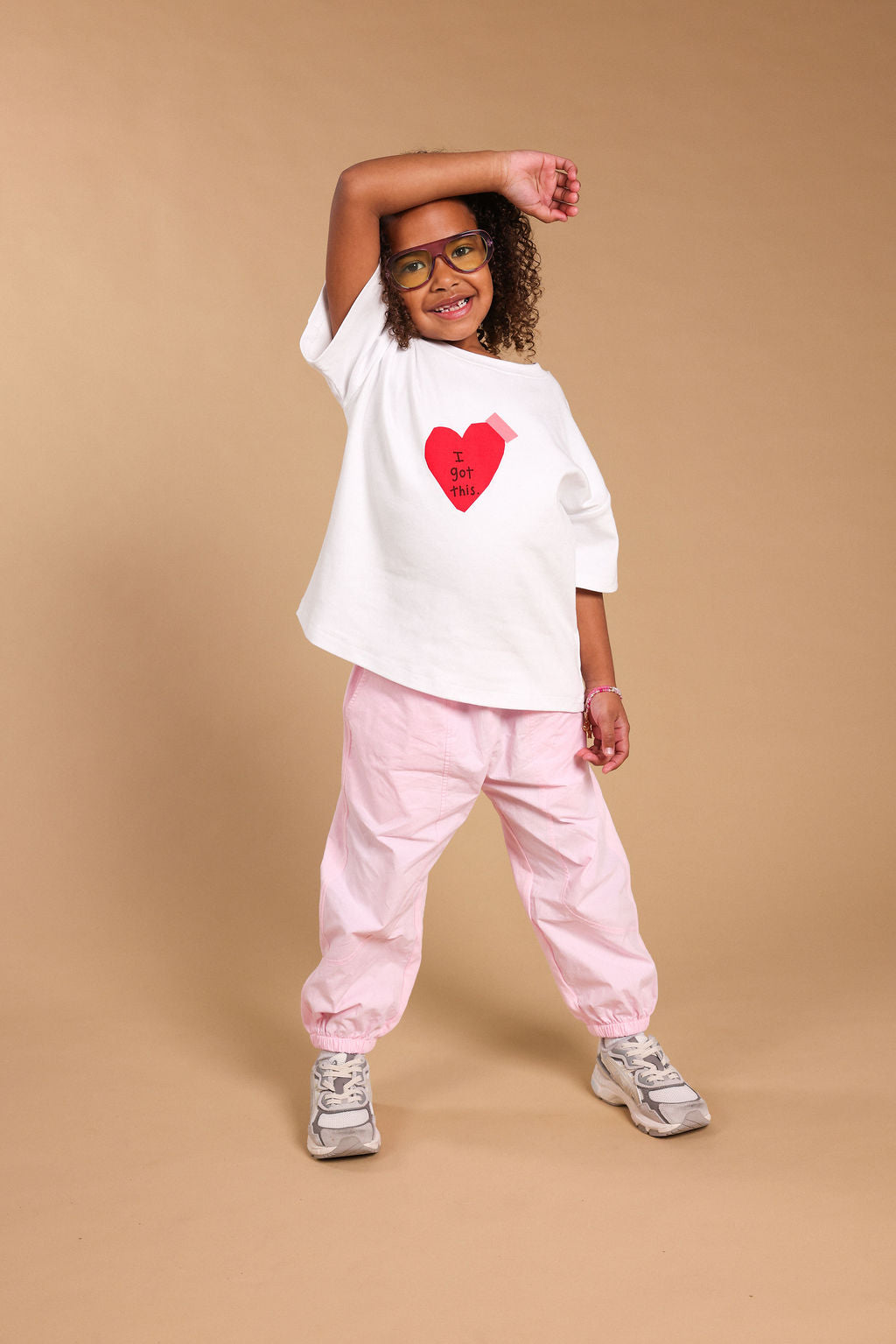 A model posing in a white oversized t-shirt with a red heart graphic 'I got this', light pink pants, and grey sneakers against a solid tan studio background