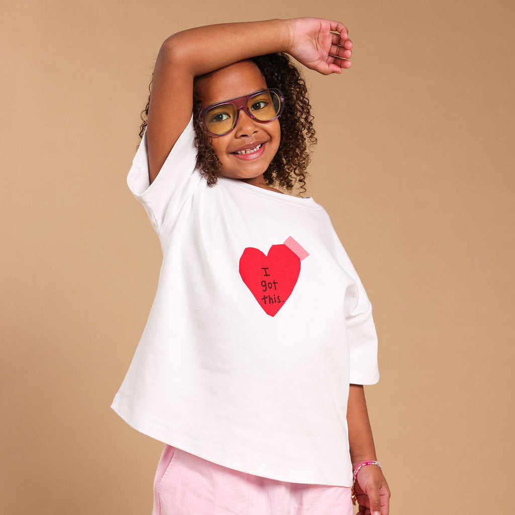 A young girl with curly hair and glasses is modeling a white oversized t-shirt with a red heart graphic that says 'I GOT YOU' in black text, paired with light pink pants, against a solid beige studio background.