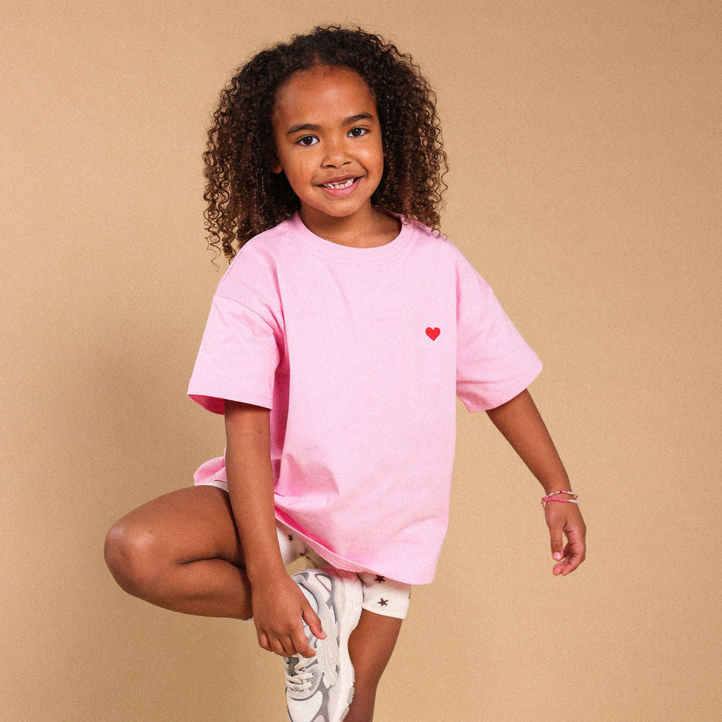 A young girl with curly hair stands in a studio wearing an oversized light pink t-shirt with a small red heart on the chest, paired with white shorts and white sneakers, against a solid beige background.
