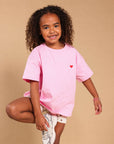 A young girl with curly hair stands in a studio wearing an oversized light pink t-shirt with a small red heart on the chest, paired with white shorts and white sneakers, against a solid beige background.