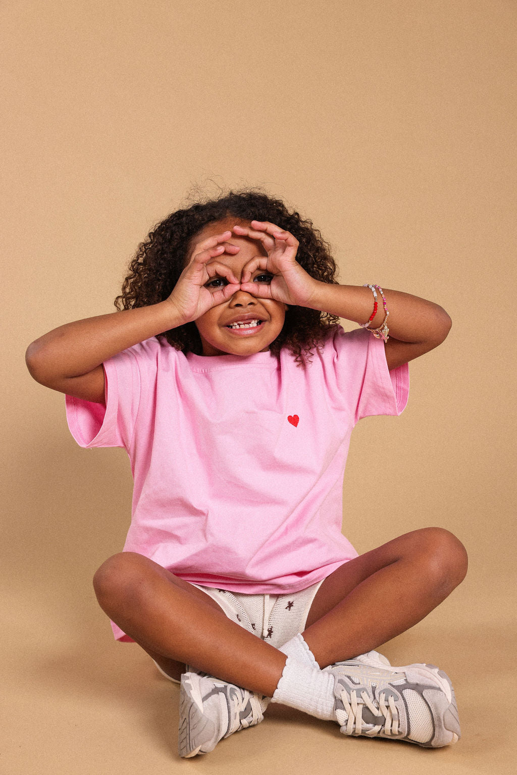 A young child with curly hair sits cross-legged against a solid beige background, wearing a pink t-shirt with a small red heart on the chest, white shorts, and white sneakers.