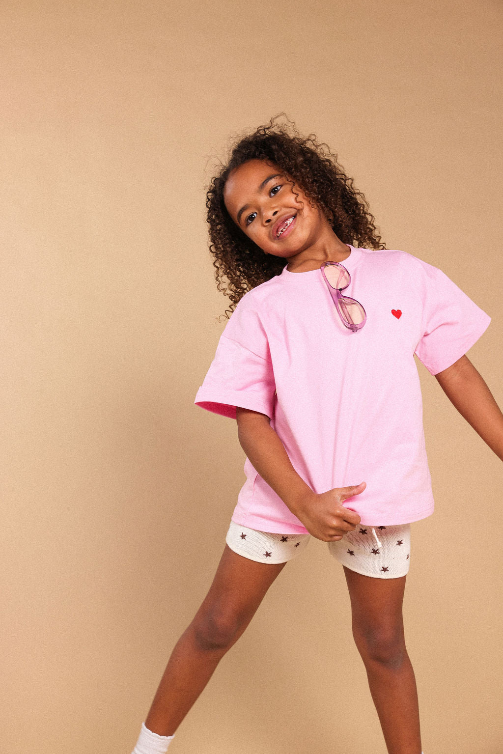 A young girl with curly hair models a light pink oversized t-shirt with a small red heart on the chest, paired with white shorts featuring a small black star pattern. She is posing against a solid beige studio background.