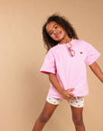 A young girl with curly hair models a light pink oversized t-shirt with a small red heart on the chest, paired with white shorts featuring a small black star pattern. She is posing against a solid beige studio background.