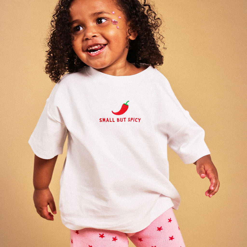 A toddler girl with curly dark hair is wearing a white oversized t-shirt with a red chili pepper graphic and the text 'SMALL BUT SPICY' on the chest, paired with pink shorts featuring a red star pattern. She is posing against a solid warm beige background.