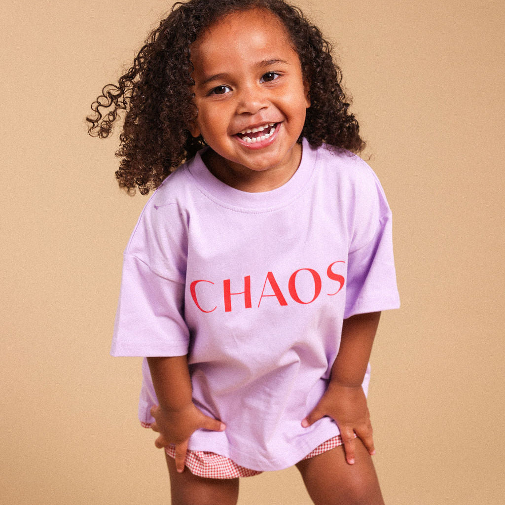A young girl with curly brown hair smiling and posing in a light purple oversized t-shirt with the word 'CHAOS' printed in red across the chest, paired with pink gingham shorts, set against a solid beige studio background.