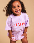 A young girl with curly brown hair smiling and posing in a light purple oversized t-shirt with the word 'CHAOS' printed in red across the chest, paired with pink gingham shorts, set against a solid beige studio background.