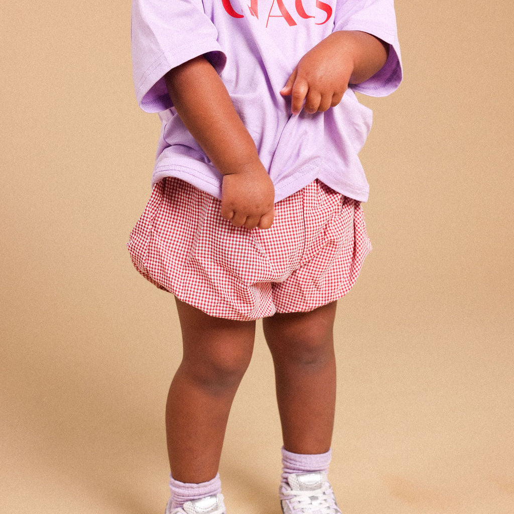 A toddler standing against a beige studio background, wearing an oversized light purple T-shirt with red text, pink and white gingham shorts, and white sneakers.