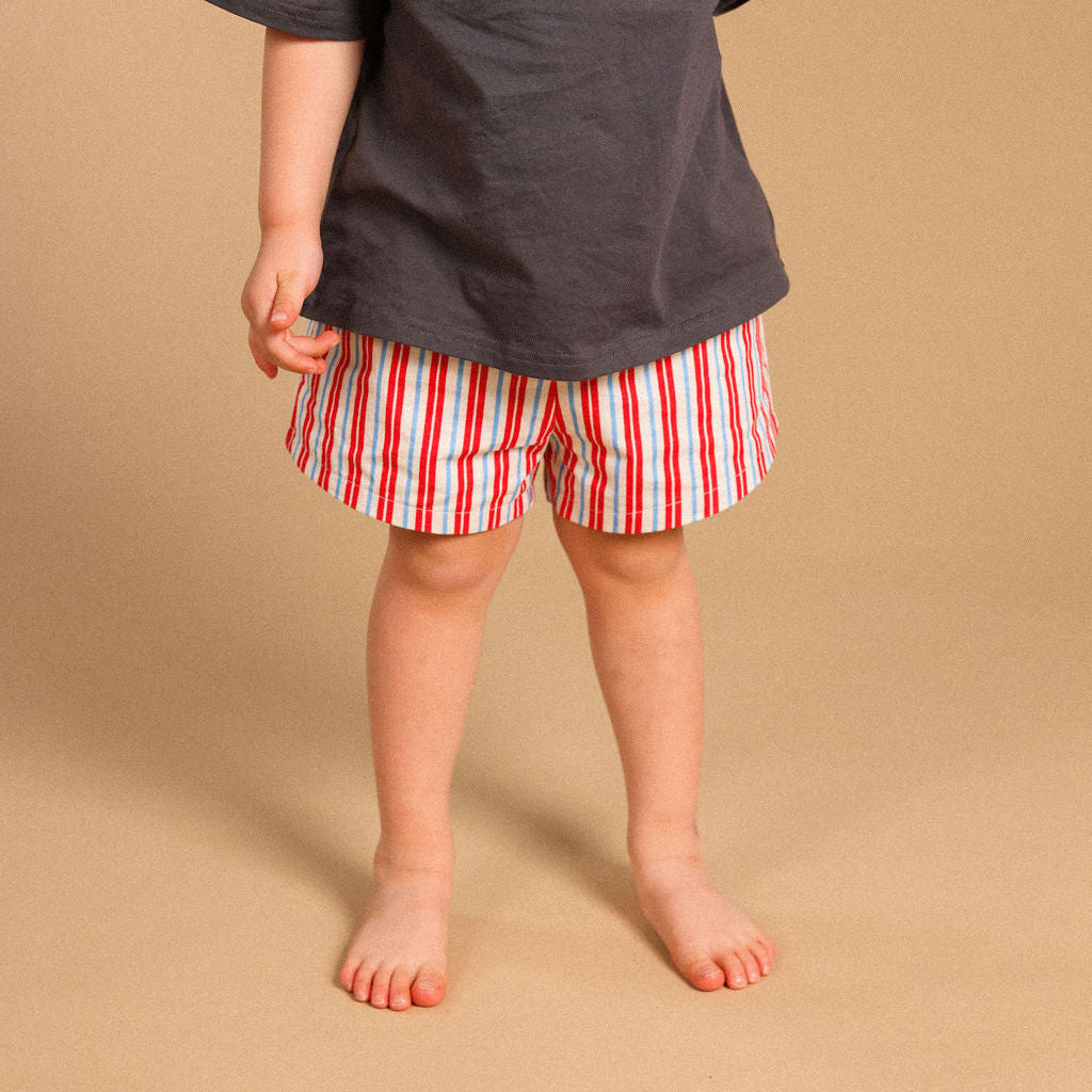 A child wearing an oversized dark charcoal gray t-shirt with the word 'CHAOS' printed in rose gold across the chest, paired with red and white vertically striped shorts, standing against a solid beige background.