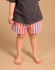 A child wearing an oversized dark charcoal gray t-shirt with the word 'CHAOS' printed in rose gold across the chest, paired with red and white vertically striped shorts, standing against a solid beige background.