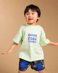 A young child modeling a light green t-shirt with blue text that reads 'DOING COOL THINGS' and blue patterned shorts, standing against a solid beige background.