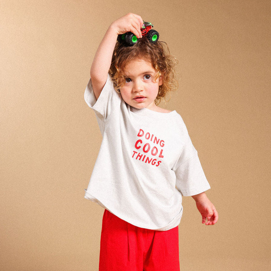 A toddler stands barefoot against a warm beige studio backdrop, wearing an oversized white T-shirt with red text reading 'DOING COOL THINGS' and bright red pants. The child is holding a small green toy car above their head.