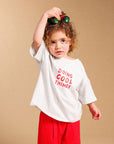 A toddler stands barefoot against a warm beige studio backdrop, wearing an oversized white T-shirt with red text reading 'DOING COOL THINGS' and bright red pants. The child is holding a small green toy car above their head.