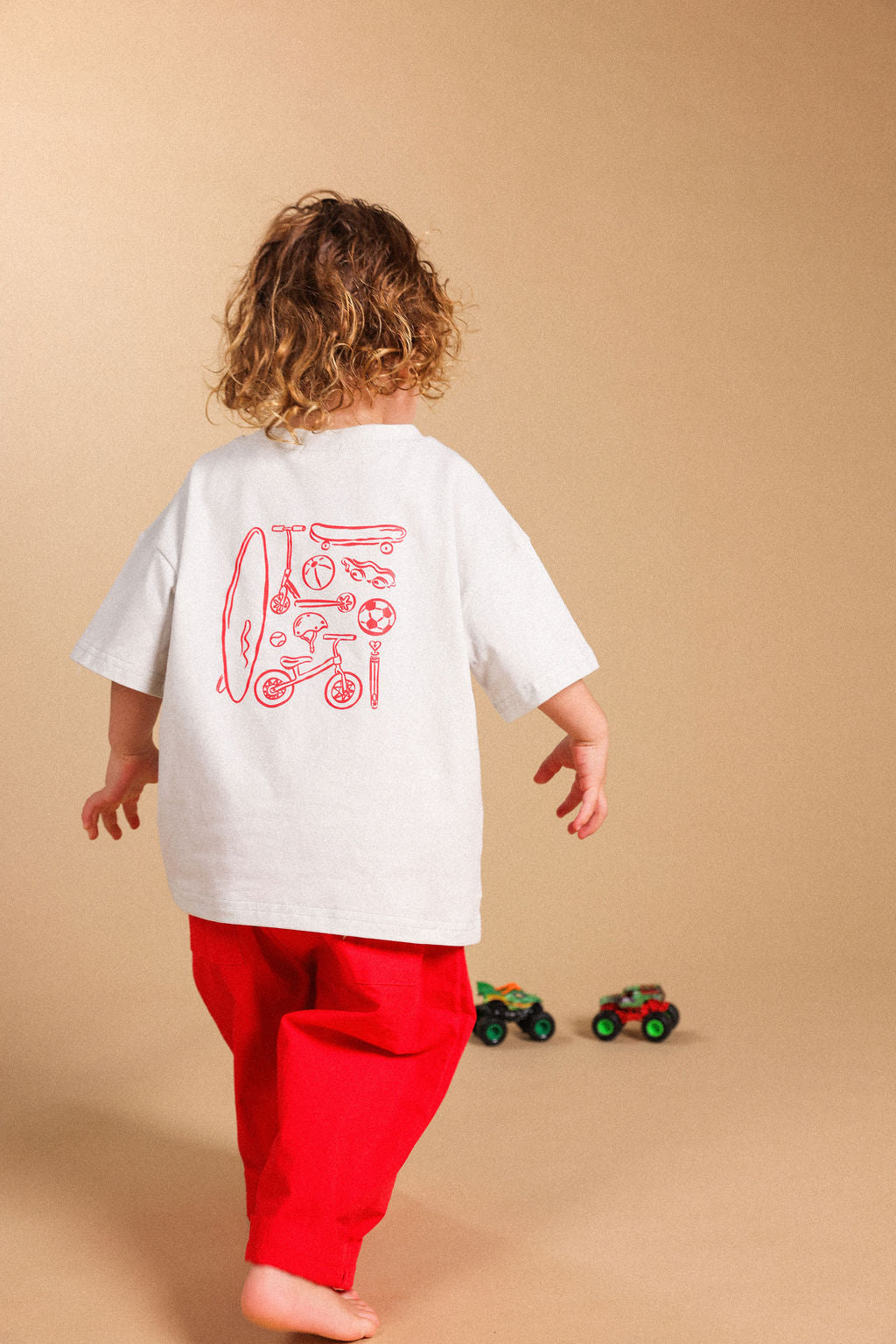 A child seen from behind wearing a white oversized T-shirt with a red graphic print on the back and bright red pants, standing against a plain beige studio background.