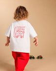 A child seen from behind wearing a white oversized T-shirt with a red graphic print on the back and bright red pants, standing against a plain beige studio background.