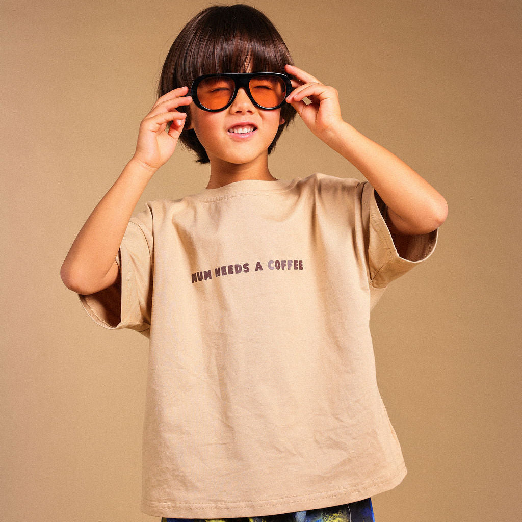 A child wearing a beige graphic t-shirt with 'MUM NEEDS A COFFEE' printed on the chest, blue and yellow patterned pants, and black-rimmed orange-tinted glasses, posing against a solid beige background.