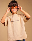 A child wearing a beige graphic t-shirt with 'MUM NEEDS A COFFEE' printed on the chest, blue and yellow patterned pants, and black-rimmed orange-tinted glasses, posing against a solid beige background.