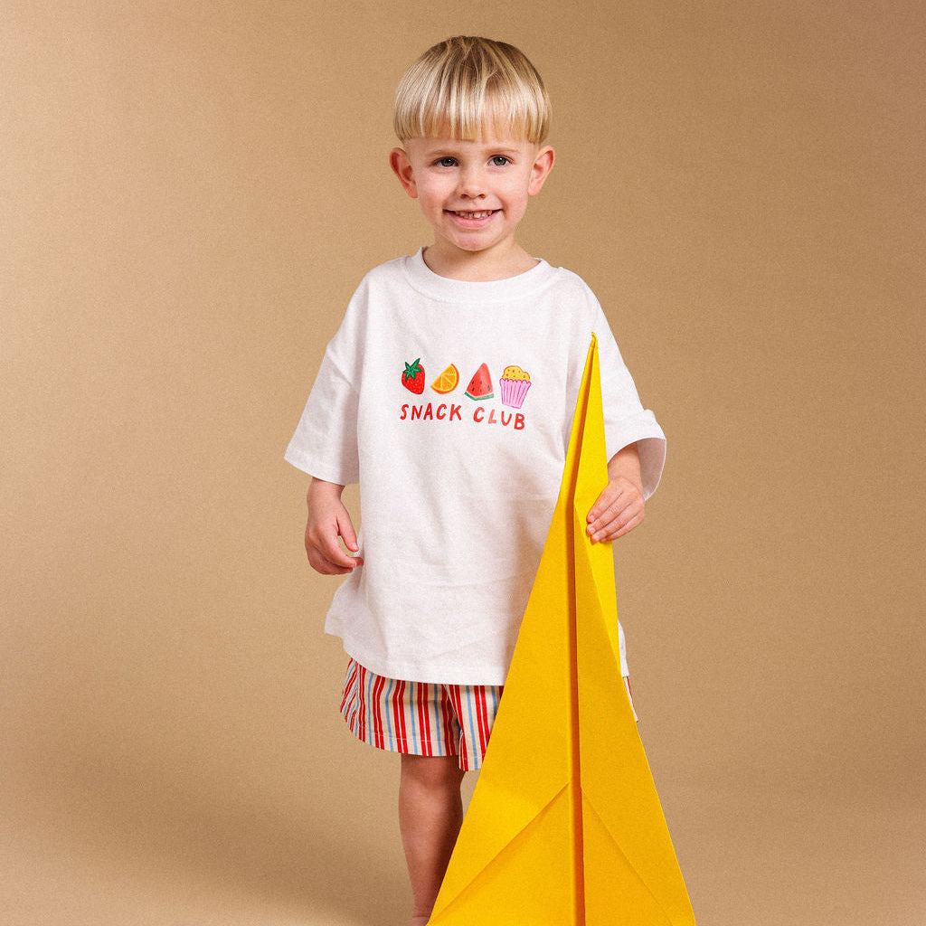 A young blonde boy stands barefoot against a solid beige studio background, wearing a white graphic t-shirt that reads 'SNACK CLUB' with colorful fruit and ice cream illustrations, red and white striped shorts, and holding a large yellow paper airplane.