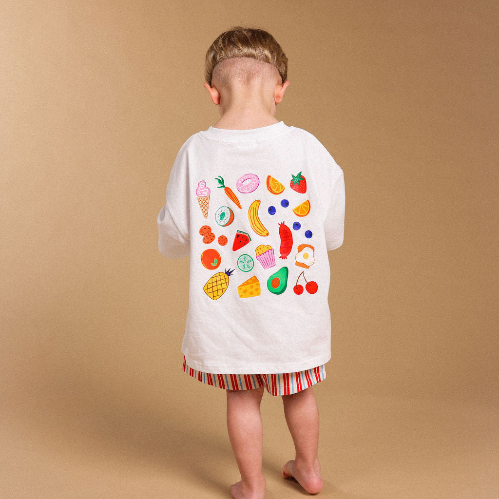 A child standing with their back to the camera, wearing a white t-shirt with a colorful fruit and vegetable pattern on the back, and red, orange, and white striped shorts, against a solid tan studio backdrop.