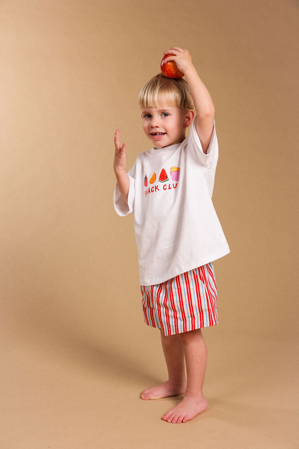 A young child stands barefoot against a plain beige studio background, holding a red apple above their head. The child is wearing a white T-shirt with a colorful graphic and the words 'SNACK CLUB' on the front, paired with red-and-white vertically striped shorts.