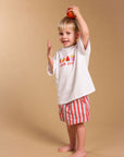 A young child stands barefoot against a plain beige studio background, holding a red apple above their head. The child is wearing a white T-shirt with a colorful graphic and the words 'SNACK CLUB' on the front, paired with red-and-white vertically striped shorts.