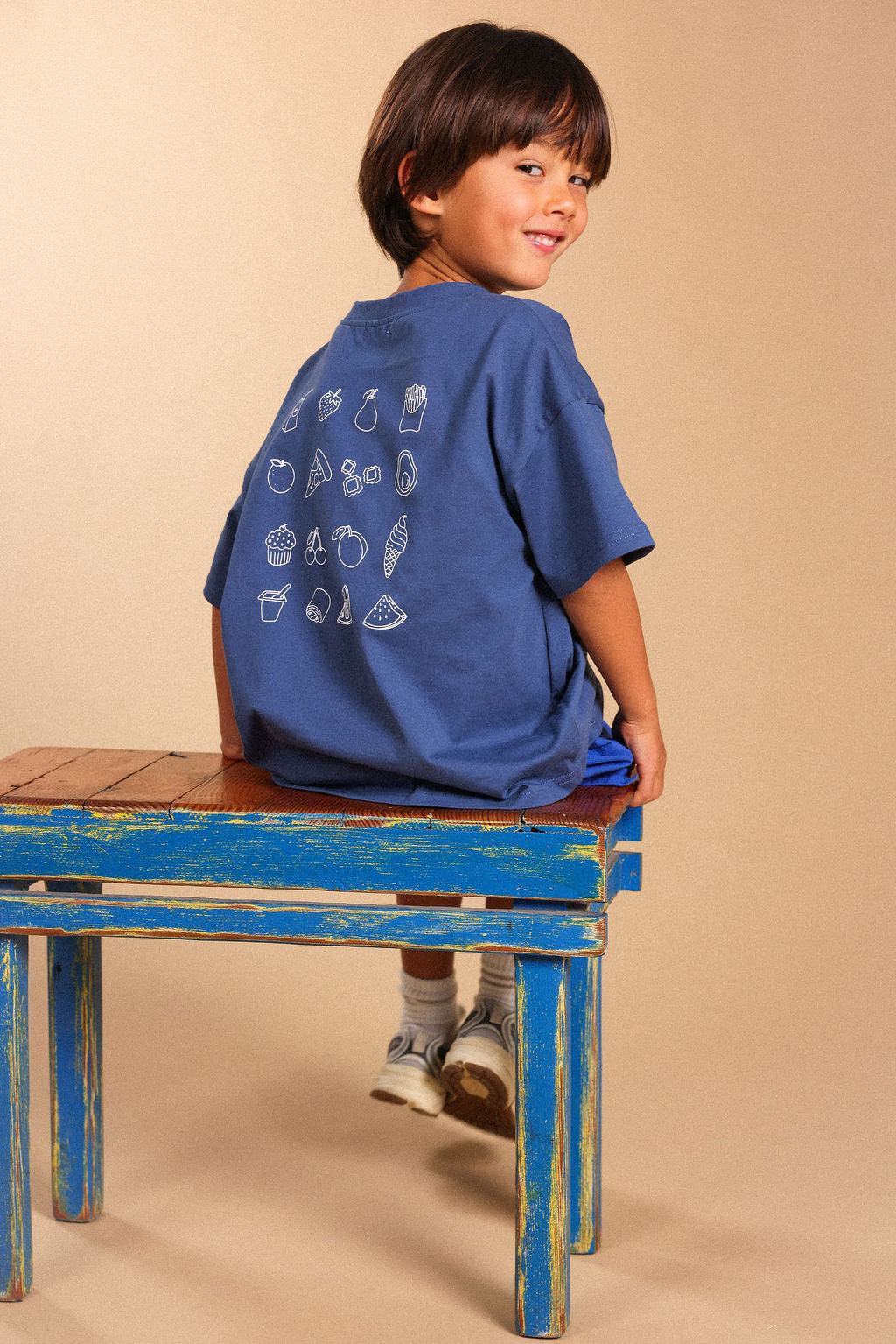 A young boy wearing a blue t-shirt with white food illustrations on the back, sitting on a rustic blue wooden bench against a solid beige background.