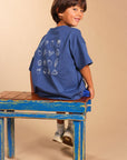 A young boy wearing a blue t-shirt with white food illustrations on the back, sitting on a rustic blue wooden bench against a solid beige background.