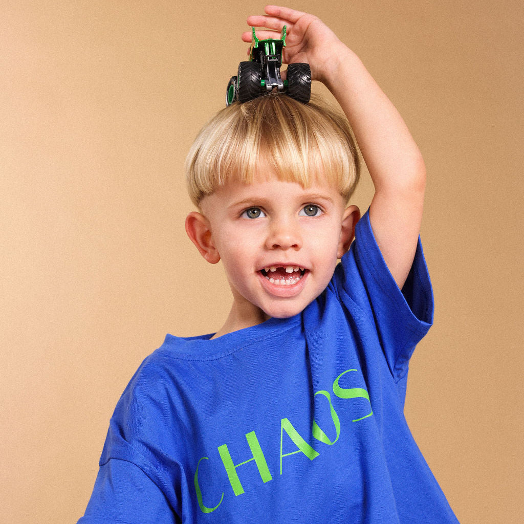 A young blonde boy wearing a blue t-shirt with CHAOS green text, balancing a green toy monster truck on his head against a solid beige background.