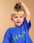A young blonde boy wearing a blue t-shirt with CHAOS green text, balancing a green toy monster truck on his head against a solid beige background.