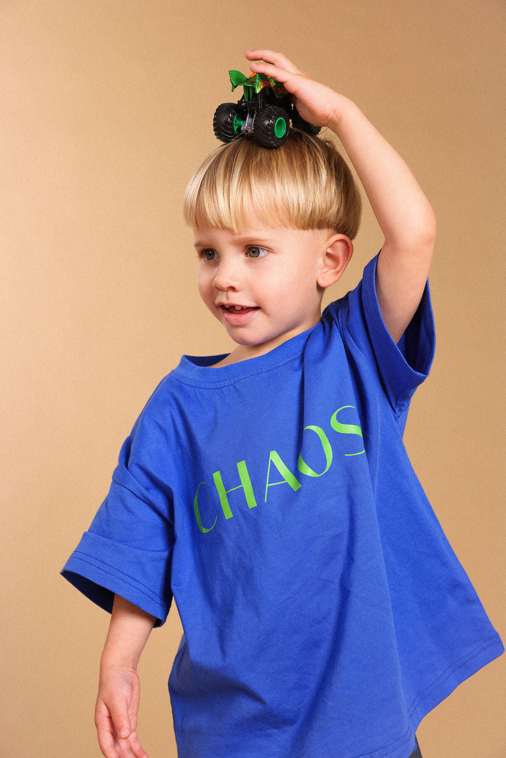 A young blonde boy wearing a blue oversized t-shirt with the word 'CHAOS' printed in green across the chest, balancing a small green toy truck on his head against a solid beige background.