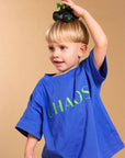 A young blonde boy wearing a blue oversized t-shirt with the word 'CHAOS' printed in green across the chest, balancing a small green toy truck on his head against a solid beige background.