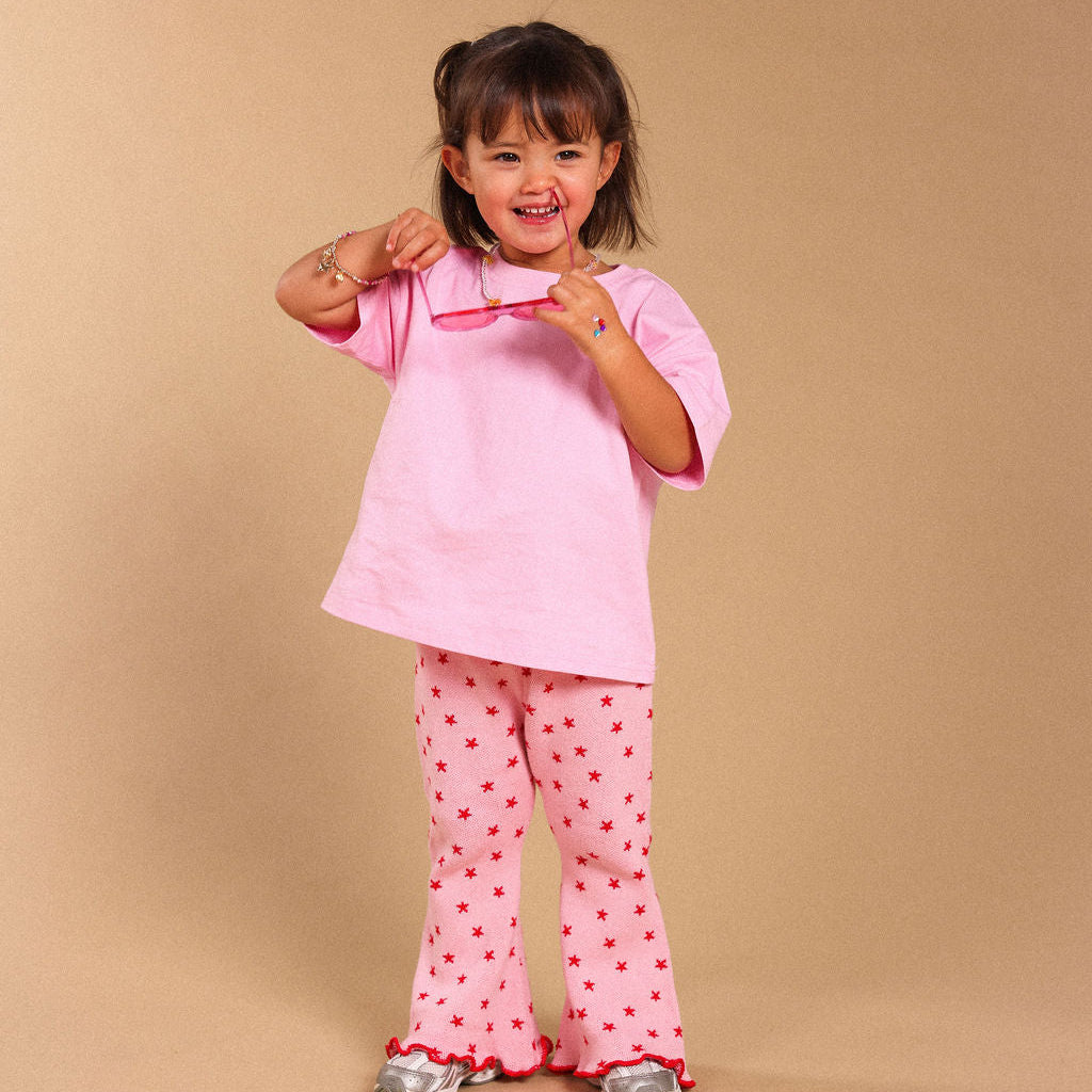 A young girl smiling and posing in a pink short-sleeve t-shirt and matching pink pants with small red star patterns, set against a solid beige studio background.