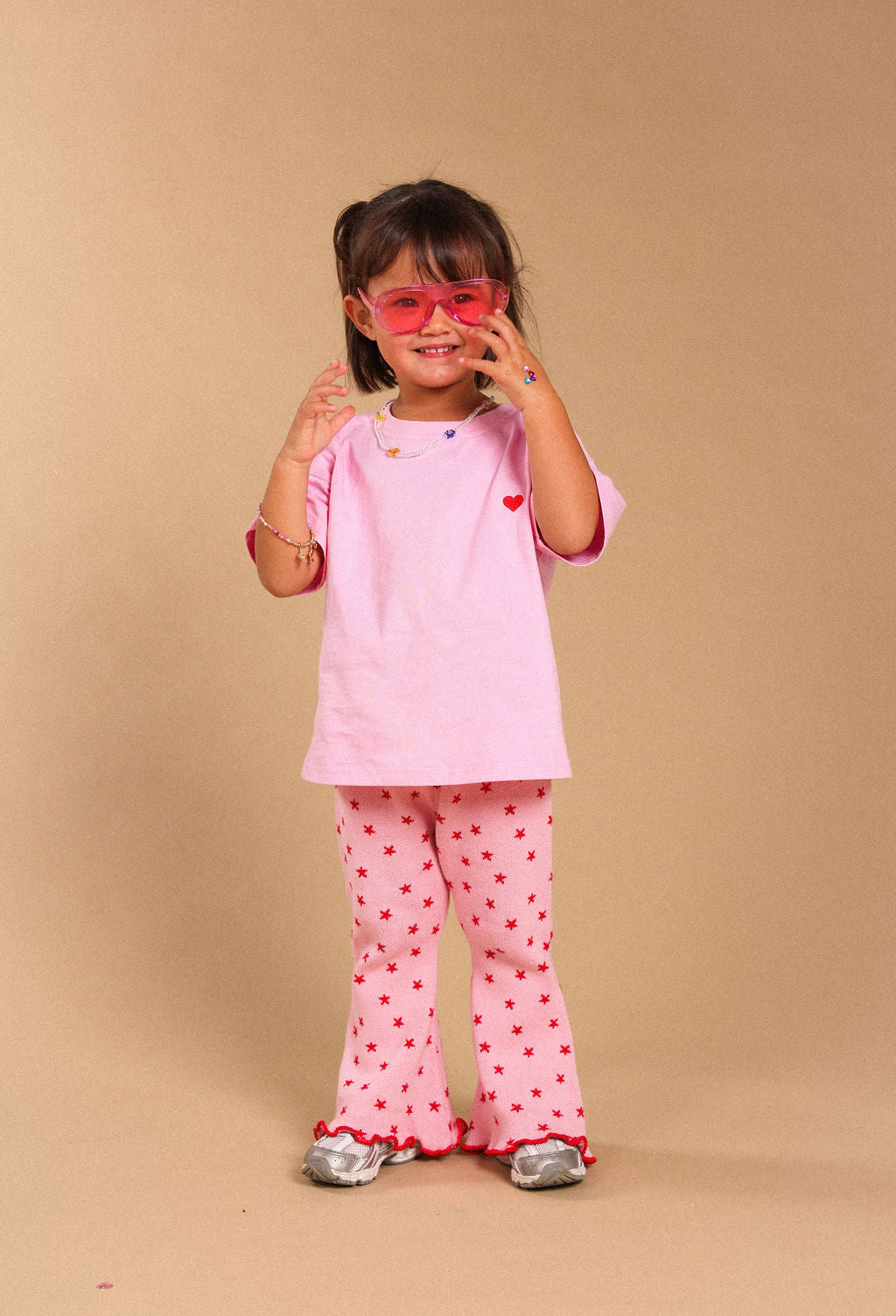 A young girl modeling a pink short-sleeve t-shirt and matching pink pants with red heart prints, accessorized with pink sunglasses, standing against a solid beige studio background.