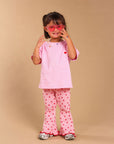 A young girl modeling a pink short-sleeve t-shirt and matching pink pants with red heart prints, accessorized with pink sunglasses, standing against a solid beige studio background.