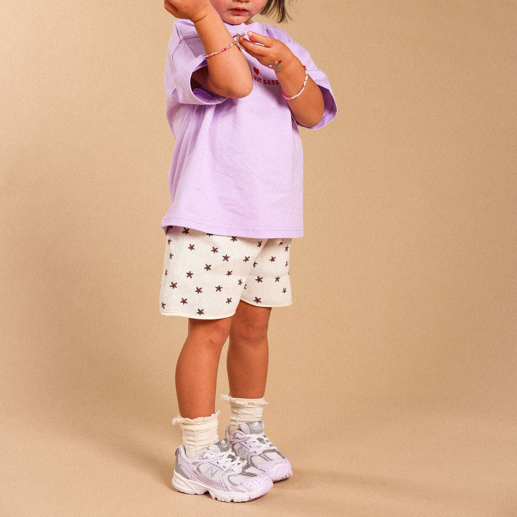 A young child wearing a light purple t-shirt, white shorts with brown star patterns, white socks, and white sneakers, standing against a solid beige studio background.