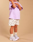 A young child wearing a light purple t-shirt, white shorts with brown star patterns, white socks, and white sneakers, standing against a solid beige studio background.