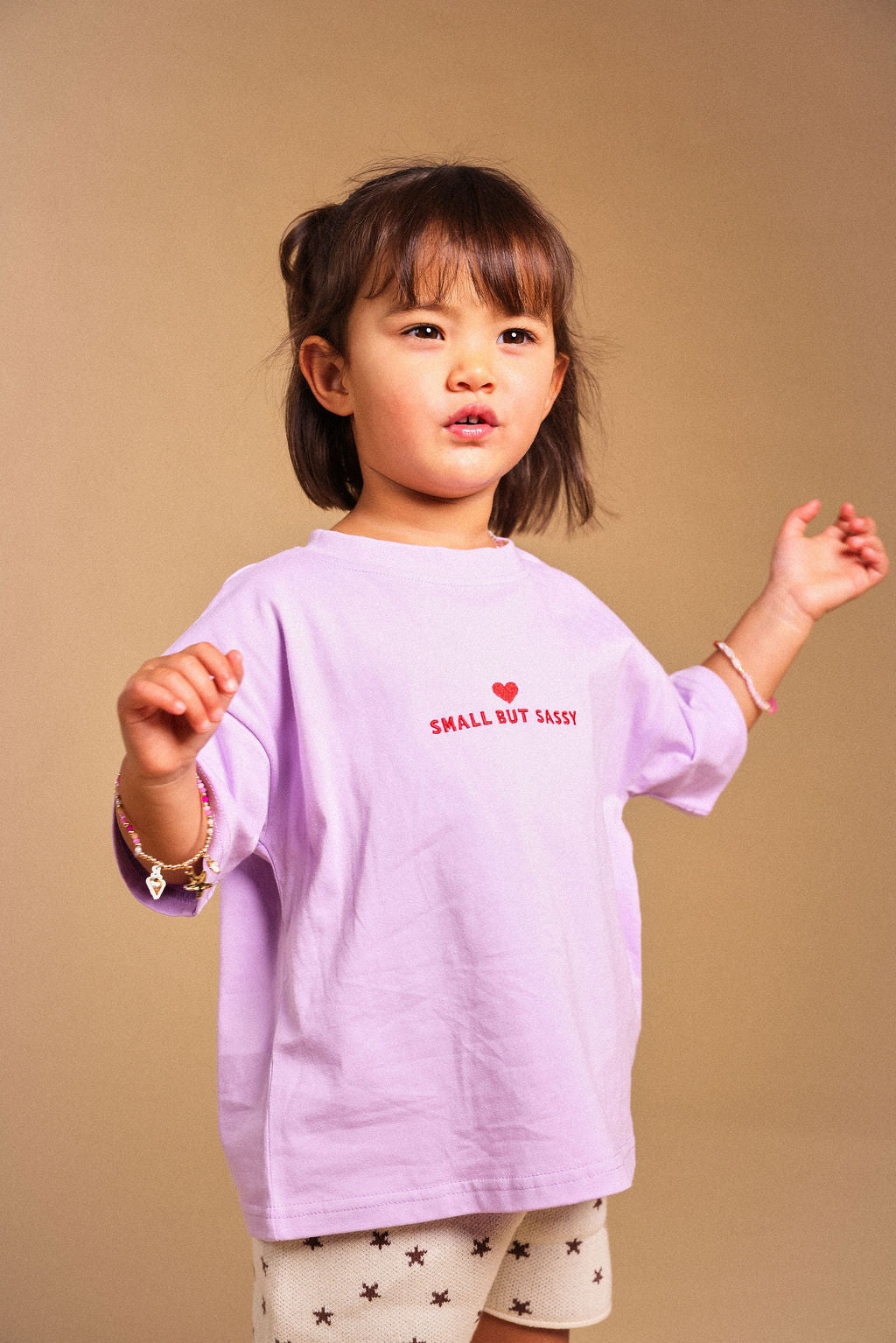 A young child wearing a light purple long-sleeve t-shirt with 'SMALL BUT SASSY' printed in red on the chest, paired with beige shorts featuring a black star pattern, set against a solid beige background.