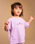 A young child wearing a light purple long-sleeve t-shirt with 'SMALL BUT SASSY' printed in red on the chest, paired with beige shorts featuring a black star pattern, set against a solid beige background.