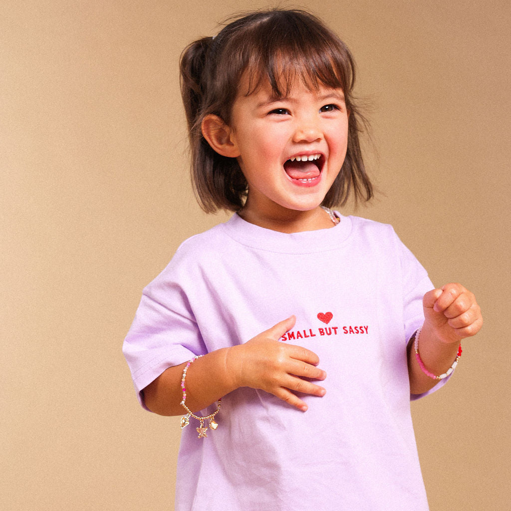 A young girl with dark hair in a ponytail is smiling and laughing while wearing a light purple t-shirt with red text that reads 'SMALL BUT SASSY' and a small red heart, paired with cream shorts decorated with small brown stars. She is standing against a solid beige studio background.