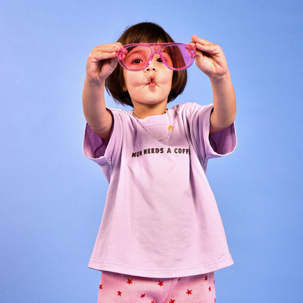 A young child wearing a light purple t-shirt with 'MUM NEEDS A COFFEE' text and pink shorts with red stars, holding pink novelty sunglasses in front of their face against a solid blue background.