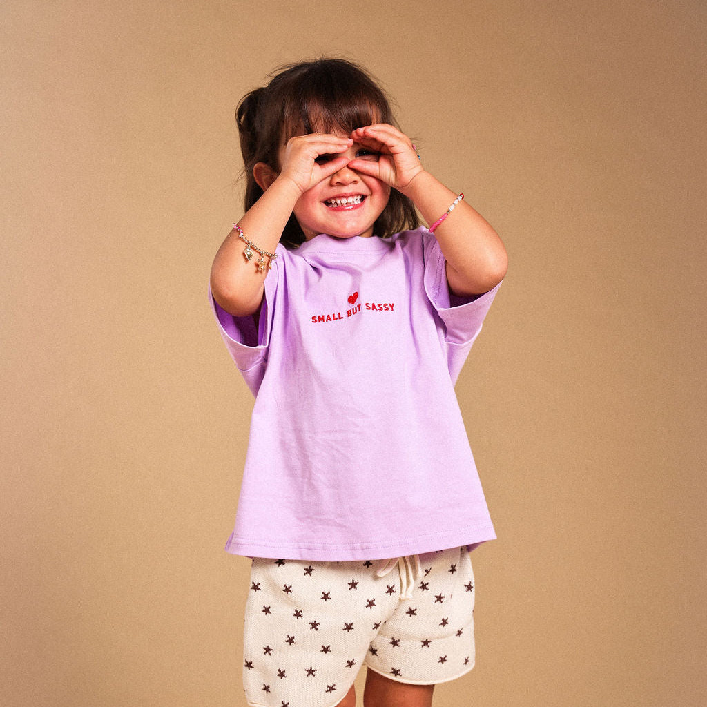 A young child modeling a light purple t-shirt with 'SMALL BUT SASSY' embroidered on the chest, paired with cream shorts featuring a brown star pattern, standing against a solid beige background.