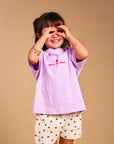 A young child modeling a light purple t-shirt with 'SMALL BUT SASSY' embroidered on the chest, paired with cream shorts featuring a brown star pattern, standing against a solid beige background.