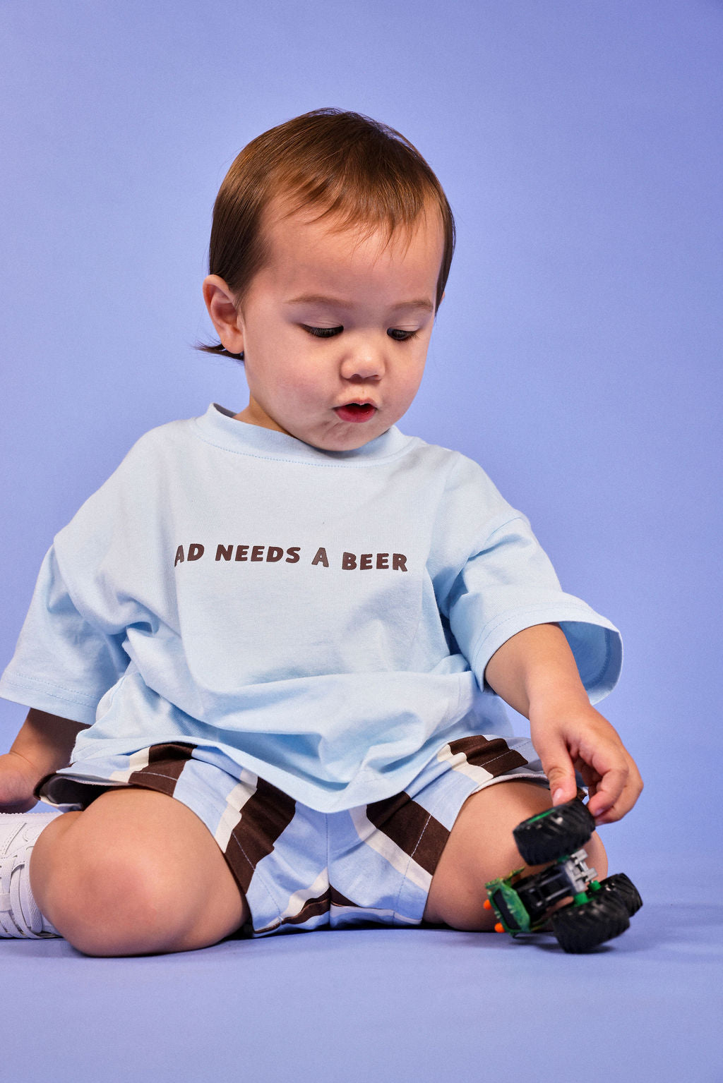 A toddler sitting on a lavender studio background, wearing a light blue t-shirt with the text 'DAD NEEDS A BEER' and brown-and-white striped shorts, holding a toy truck.
