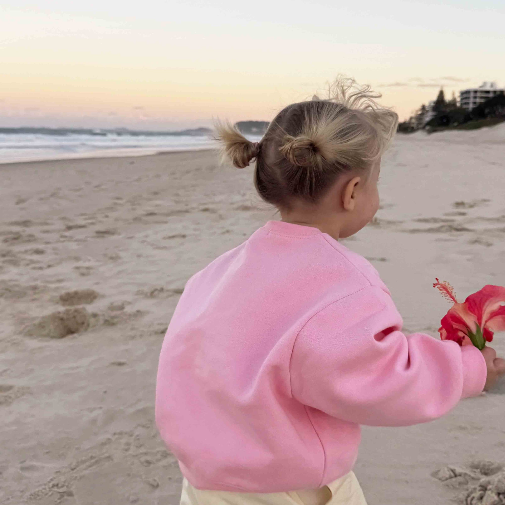 A toddler standing on a sandy beach at sunset, wearing a pink sweatshirt and cream pants, holding a red flower.