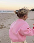 A toddler standing on a sandy beach at sunset, wearing a pink sweatshirt and cream pants, holding a red flower.