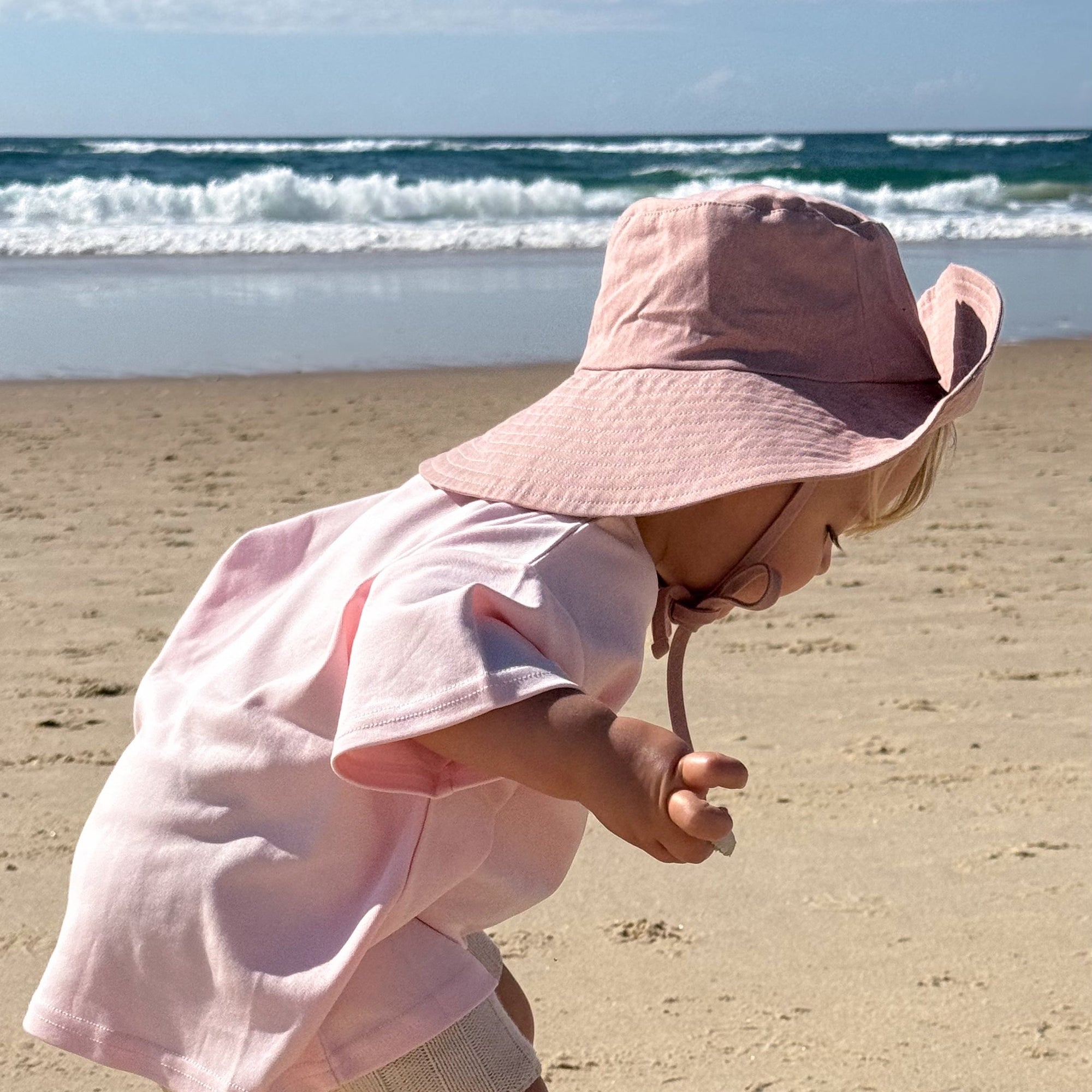 A toddler wearing a light pink sun hat and matching pink shirt stands barefoot on a sandy beach, looking down toward the sand with the ocean and blue sky in the background.